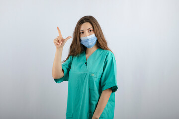 Young beautiful nurse in green uniform in a medical mask pointing up