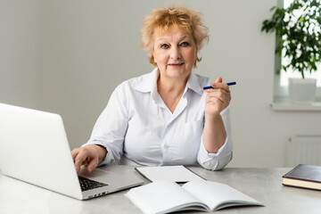 Senior woman at home in front of her laptop making notes during watching an online english language lesson by a female teacher, e-learning concept.
