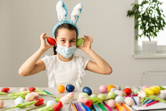 Little girl in a medical mask paints Easter eggs with a pattern. Stay home.