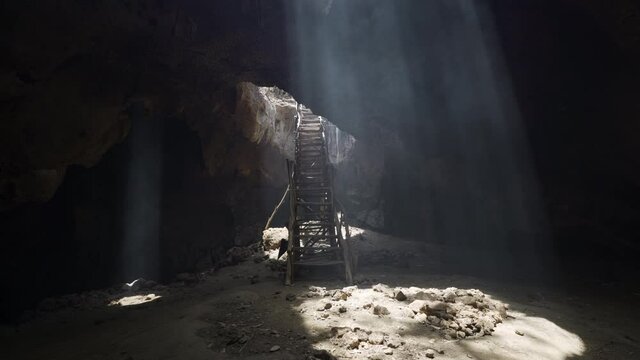 Rickety wooden stairs stretch into a dark stone cave with sun rays illuminating a sandy floor - Lombok, Indonesia