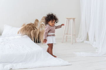 Little curly-haired African American girl plays, runs by the white bed, has fun, laughs, smiles....