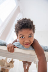 Fototapeta premium Little African American boy has fun, lies on white wooden chair, flounders with his feet. Minimalist interior, pampas grass, driftwood on the wall, airy doors on the windows. Scandinavian style.