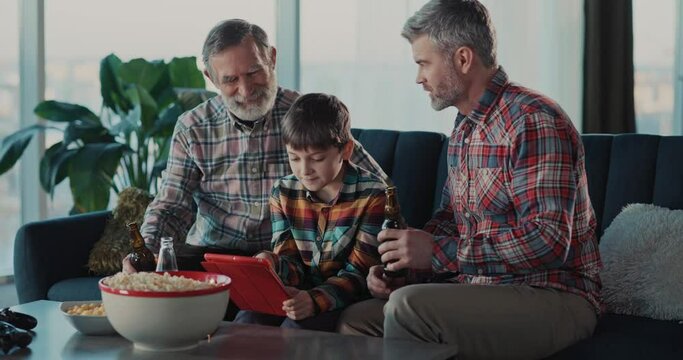 Young teenager boy using tablet computer playing games spending time with family. His father and grandpa drinking beer alcohol resting at home apartment.