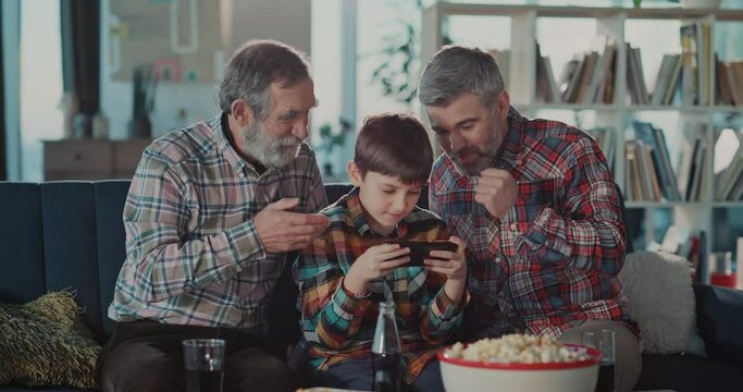 Young teenager boy playing computer game mobil ephone getting much support from his father and grandpa spending leisure time together.