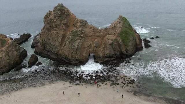 Aerial shot of people at beach with famous Keyhole Arch, drone descending forward towards waves splashing on shore - Big Sur, California