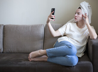 a young woman in a white T-shirt and a towel on her head with a cosmetic moisturizing mask sits on the couch and takes a selfie