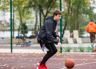 Fototapeta premium Cute Teenager with orange basketball ball plays basketball on street playground in spring summer. Hobby, active lifestyle, sports activity for kids.