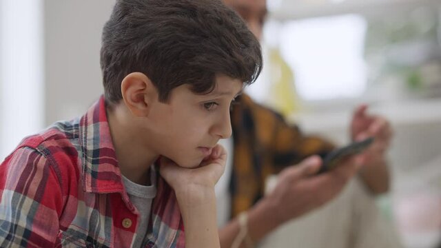 Close-up of sad charming Middle Eastern kid sitting indoors with blurred angry brother arguing at background gesturing and pointing at smartphone. Headshot of upset boy scolded by sibling.