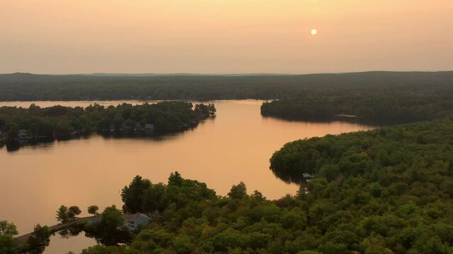 Aerial panning shot of lake with houses amidst green trees against sky, drone flying over landscape at sunset - Berkshire County, Massachusetts