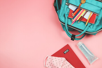 Top view of red 2021 diary, backpack with school supplies , pink polka dots fabric mask and alcohol sanitizer gel on pink background. COVID-19 prevention while going back  to school in 2021