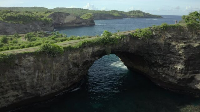 Aerial moving quickly forward to a natural rock arch bridge formation spanning a beautiful ocean bay with a mona standing on the edge of the cliff admiring the view - Nusa Penida & Lombok, Bali