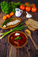 Borscht with sour cream in a wooden bowl on a wooden table. The table also contains black bread, bacon, green onions, peppers and garlic. Traditional Ukrainian and Russian cuisine.