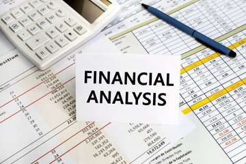 Naklejka na ściany i meble The inscription financial analysis on a white sheet that stands on a financial document on the background of a white calculator and pen. Business and financial concept
