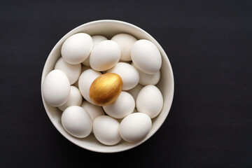 Golden egg lies on top of white ones in a bowl on black background. Easter or wealth concept. Minimalist
