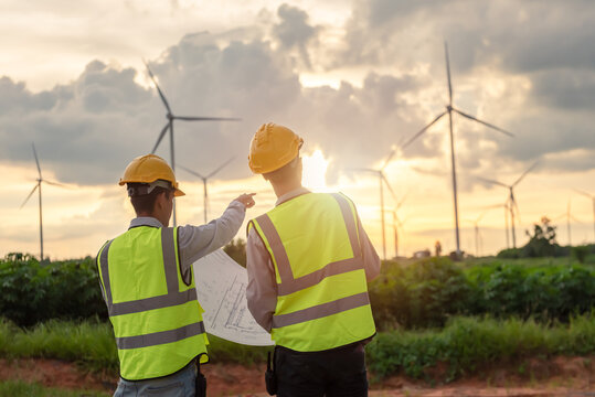 two asian male engineers working at turbines farm during beautiful sunset. Renewable energy technology and sustainability. Alternative energy for future