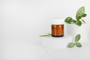 Herbal pills in glass bottle and green plant leaves on podium on white background. alternative...