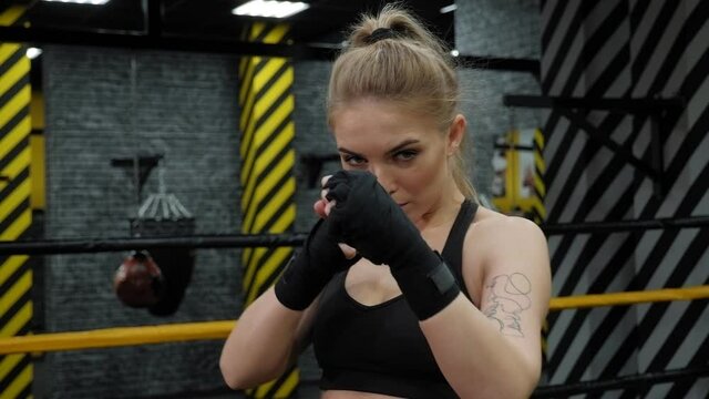 Portrait of a young female kickboxer in the gym with black bandages on her hands, she stands in a boxing stance in the ring.