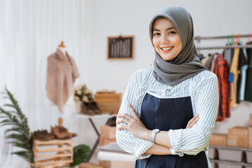 portrait of young attractive muslim woman standing with arm crossed in a room with desk,mannequin,and clothing in designer workspace