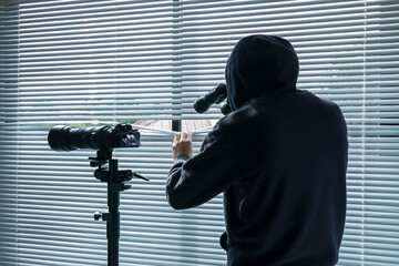 The man in black looks binoculars through the blinds.