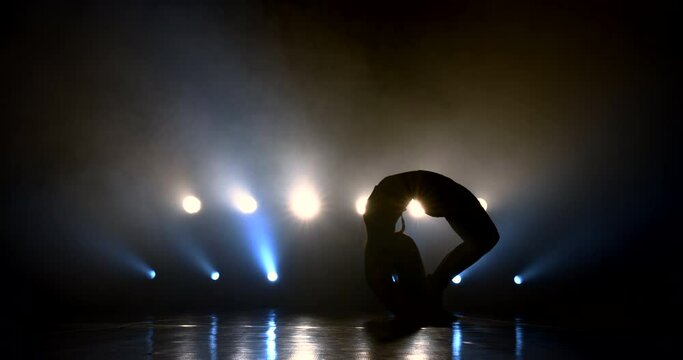 The silhouette of the dancer flaps her hair, gracefully makes a back somersault and erotically lies on her stomach under the bright spotlights.