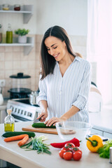 Obraz na płótnie Canvas Cute happy young brunette woman in good mood preparing a fresh vegan salad for a healthy life in the kitchen of her home