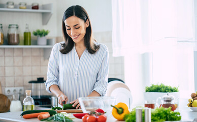 Cute happy young brunette woman in good mood preparing a fresh vegan salad for a healthy life in the kitchen of her home