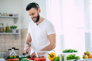 Handsome smiling young bearded man cooks fresh healthy vegan salad for his family and having fun in...