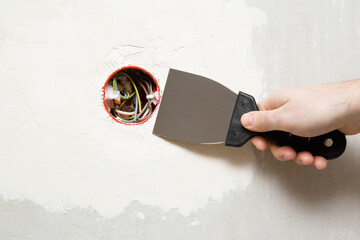 Young adult man hand using spatula and plastering wall with white fresh putty around new plastic outlet. Closeup. Repair work of home. Front view.