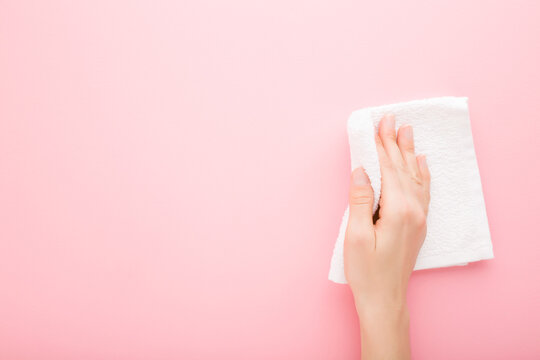 Young adult woman hand holding white rag and wiping table, wall or floor surface in kitchen, bathroom or other room. Empty place for text or logo. Light pink background. Pastel color. Top down view.