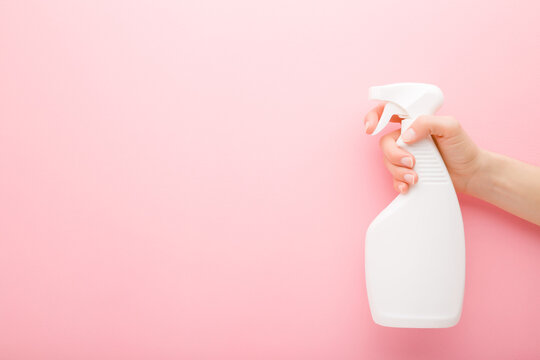 Young adult female hand holding white spray bottle. Detergent for different surfaces in kitchen, bathroom and other rooms. Closeup. Empty place for text or logo. Light pink background. Pastel color.