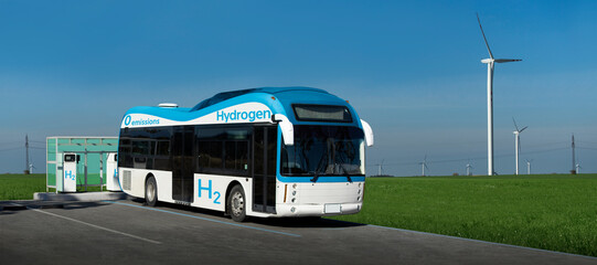 A hydrogen fuel cell bus stands at the filling station on a background of wind turbines
