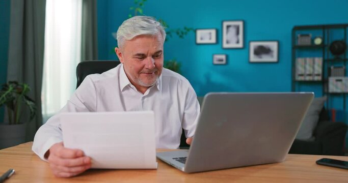 Happy serious old gray-haired mature male reading medical report, working with paper documents sitting at desk in office room in home. Tracking shot in slow motion.