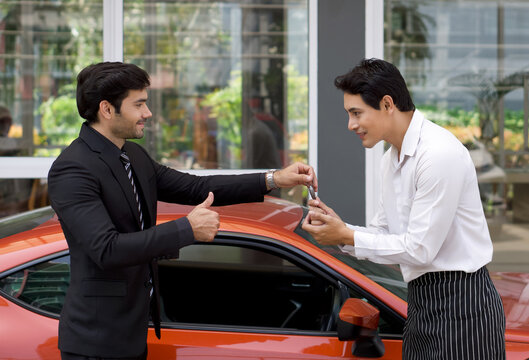 A Persian businessman in a black suit smiles and thumbs up while handling sport car key to the valet service staff.