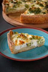 A piece of traditional french open quiche pie with feta cheese and broccoli on a dark green background.