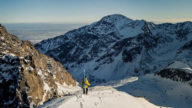 Adult man stand on the mountain top cliff with backpack and ski observe long alpine panorama to concur skiing. Hiking adventure lifestyle extreme vacations.