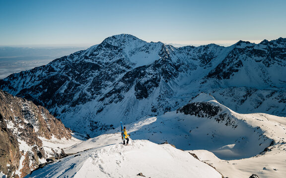 Adult man stand on the mountain top cliff with backpack and ski observe long alpine panorama to concur skiing. Hiking adventure lifestyle extreme vacations.