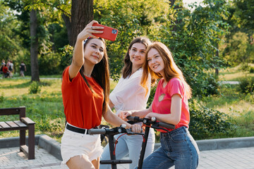 Obraz premium Three young girl friends on the electro scooters taking picture of themselves on cell phone at summer sunny day. Outdoor portrait of three friends girl riding electric kick scooter in the park