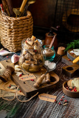 homemade marinated mushrooms in glass jar on wooden board with herbs, dill. pepper, vintage spoons