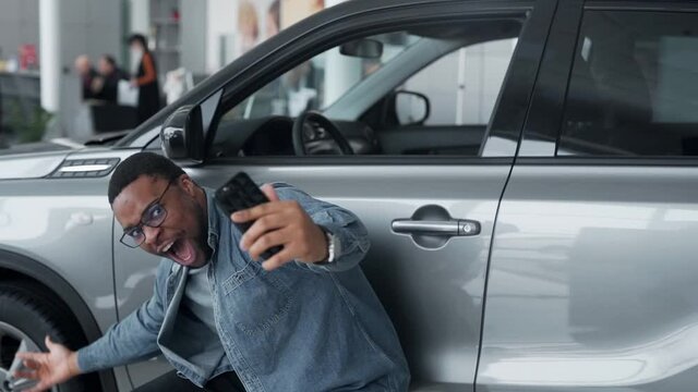 A black man happily shoots a video near the car in a car dealership