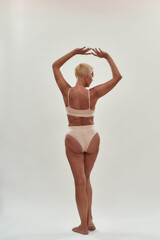 Feeling sexy at any age. Rear view of a mature woman in underwear keeping arms over the head while posing half naked against light background