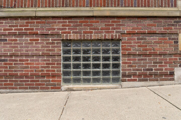 Straight on view of a glass block basement window in an old red brick building, sloping sidewalk foreground, horizontal aspect