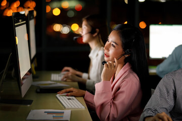 Portrait adult asian woman who is call center wearing headphone looking at screen monitor and taking phone typing on keyboard and graph chart in paper not far at her in night bokeh on blur background