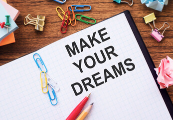 Notepad with the text MAKE YOUR DREAMS on the table with colored paper clips and crumpled sheets.