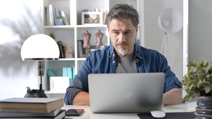 50s businessman with gray hair working from home. Man in casual sitting at desk using laptop computer, business manager online in home office.