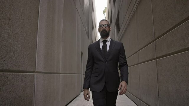 Young Man In Business Clothes Walks Down Narrow Street Between Two Buildings. Stylish Young Man In Suit With Beard And Glasses.