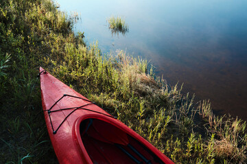 An empty red kayak by the water, on the green shore bathed in the dawn sun. 