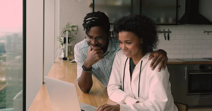Happy young bonding african american family couple looking at laptop screen, planning vacation, booking hotel online, web surfing information or shopping together, sitting at table in modern kitchen.