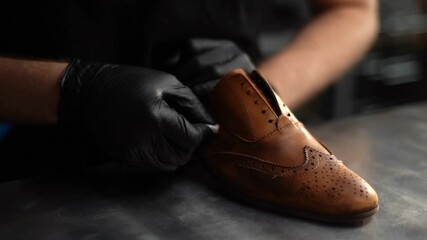 Close-up of unrecognizable shoemaker wearing black latex gloves polishing old light brown leather shoes. Process of repairing and restoration shoes by cobbler in shoe repair shop with dark interior.