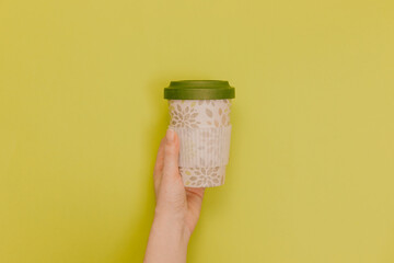  Hand holding bamboo reusable cup with lid on. Reusable tea cup on a yellow background. Zero waste. A reusable green thermos Cup on a yellow background. The concept of zero waste