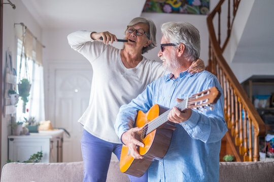 Couple of two happy seniors or mature and old people singing and dancing together at home indoor. Retired man playing the guitar while his wife is singing with a remote control of TV.